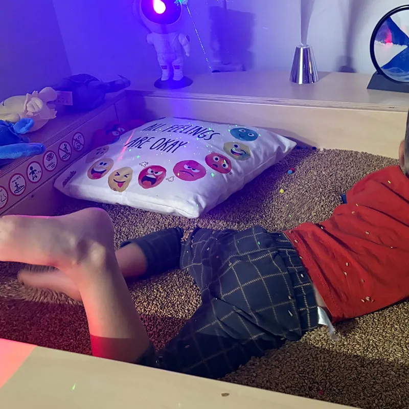 Mājīgā, rotaļīgā iekštelpu vidē bērns guļ sensorajā kastē, kas piepildīta ar maziem brūniem siltiem graudiem, blakus baltam spilvenam, kas rotāts ar krāsainām emocijzīmju sejām un uzrakstu angļu valodā ALL FEELINGS ARE OKAY.