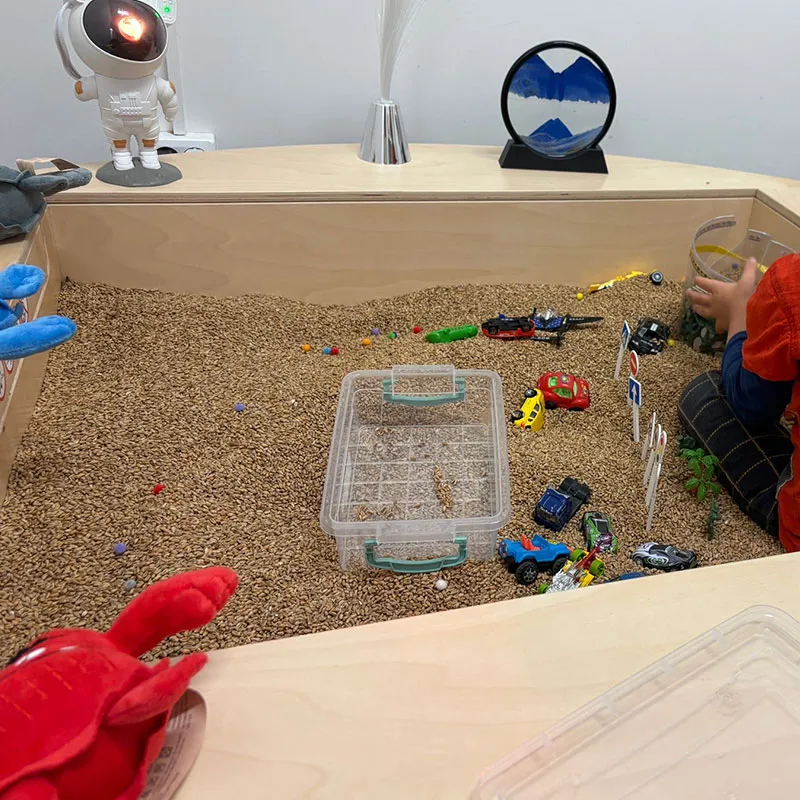 Bērns spēlējas koka sensorajā silto graudu kastē, kas piepildīta ar maziem brūniem graudiem, rotaļu automašīnām un figūriņām. Uz apkārtējā galda atrodas caurspīdīgs plastmasas trauks, astronauta lampa un dekoratīvi priekšmeti.