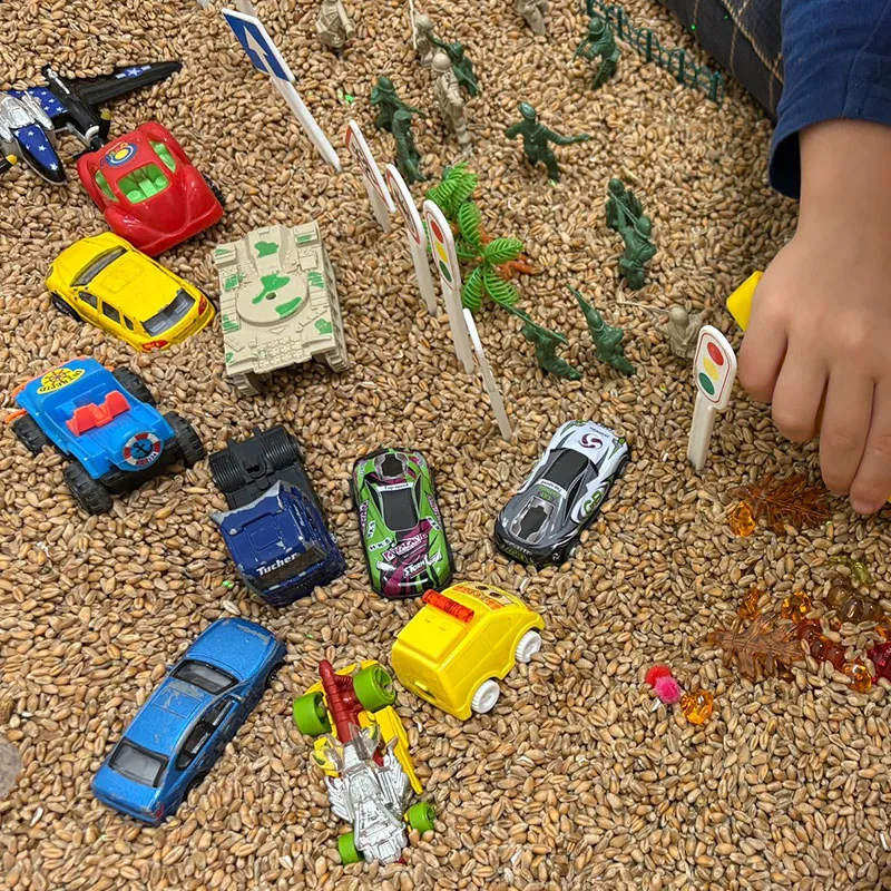 Bērna roka spēlējas ar rotaļu automašīnām, ceļa zīmēm, plastmasas dinozauriem un karavīriem, kas izkārtoti uz virsmas, kas klāta ar maziem brūniem graudiem. Krāsainie auto un rotaļlietas rada rotaļīgu ainu.